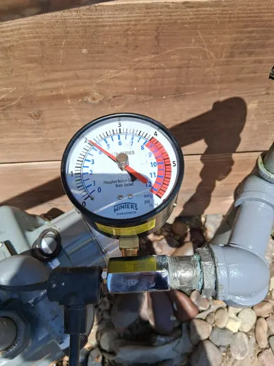 A pressure gauge attached to an outdoor gas valve near a wood fence for a leak check.