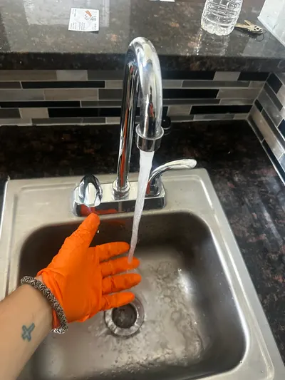 A gloved hand tests water flow from a kitchen faucet over a stainless steel sink.