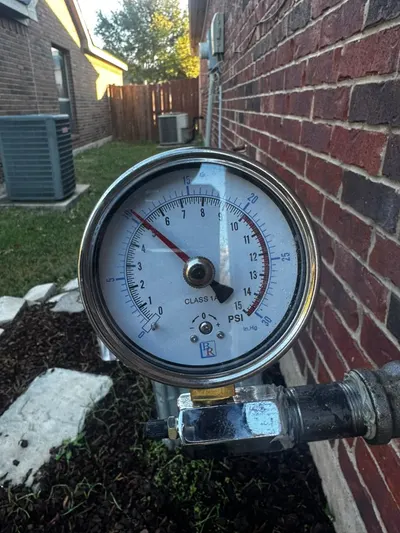 A pressure gauge attached to an exterior hose bib reads water pressure against a brick wall.