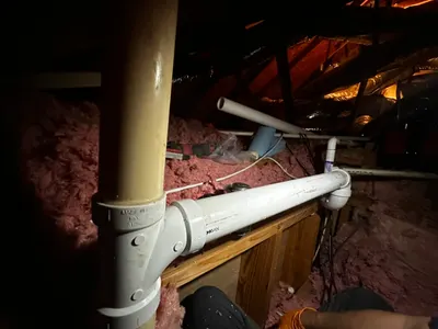 PVC vent piping routed through attic insulation and roof framing during a residential plumbing job.