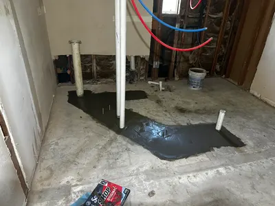 Fresh gray concrete patches a slab repair with new PEX supply lines exposed on the wall behind.