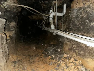 A dark view down a dug tunnel toward a suspended pipe fitting beneath a house.
