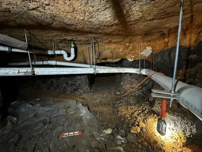 A work light illuminates a run of suspended PVC pipe inside an underground foundation tunnel.