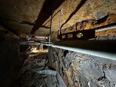 A long hand-dug tunnel beneath a house with new pipe suspended and leveled along its length.