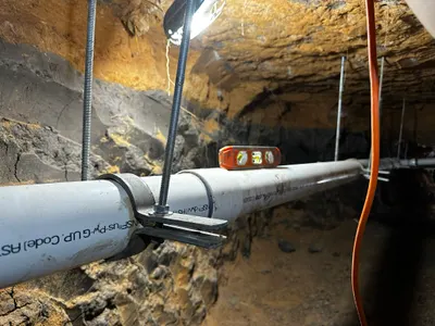 A level rests on a strapped pipe run being checked for slope inside a foundation tunnel.