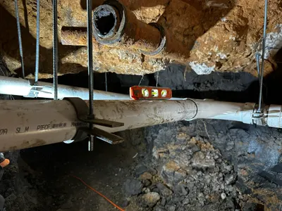 New PVC pipe strapped and leveled beneath an old rusted pipe stub in a dug foundation tunnel.