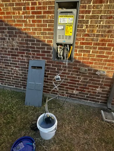 An exterior tankless water heater mounted on a brick wall with hoses draining into a bucket.