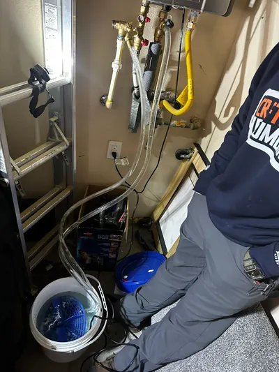 A Craftsman Plumbing technician flushes a water heater through clear hoses into a bucket in a utility closet.